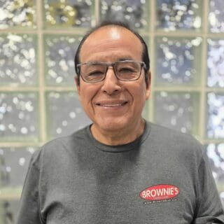 Alejandro Mancilla, machinist at Brownie's THIRD LUNG, smiling in a gray shirt with the Brownie’s logo, showcasing expertise in precision and craftsmanship for diving equipment.