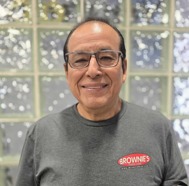 Alejandro, Machinist at Brownie's THIRD LUNG, smiling in a gray Brownie's branded shirt.
