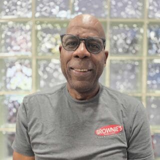 Herb Green, technician at Brownie's THIRD LUNG™, smiling in a gray shirt with the brand logo, in front of a textured glass background, showcasing his role in service and repair of dive systems.