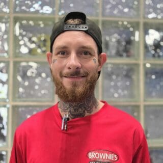 Michael Morrow, technician at Brownie's THIRD LUNG™, wearing a red shirt with logo, smiling in front of glass block background, representing expertise in diving equipment repairs and service.