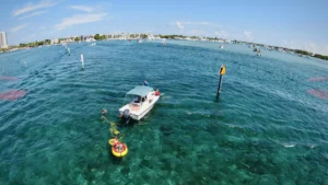 A diver uses a Brownie’s Third Lung tankless dive system connected to a small boat anchored in clear blue water near a channel marker, with the yellow and red floating compressor visible on the surface and other boats in the distance under a bright, sunny sky.