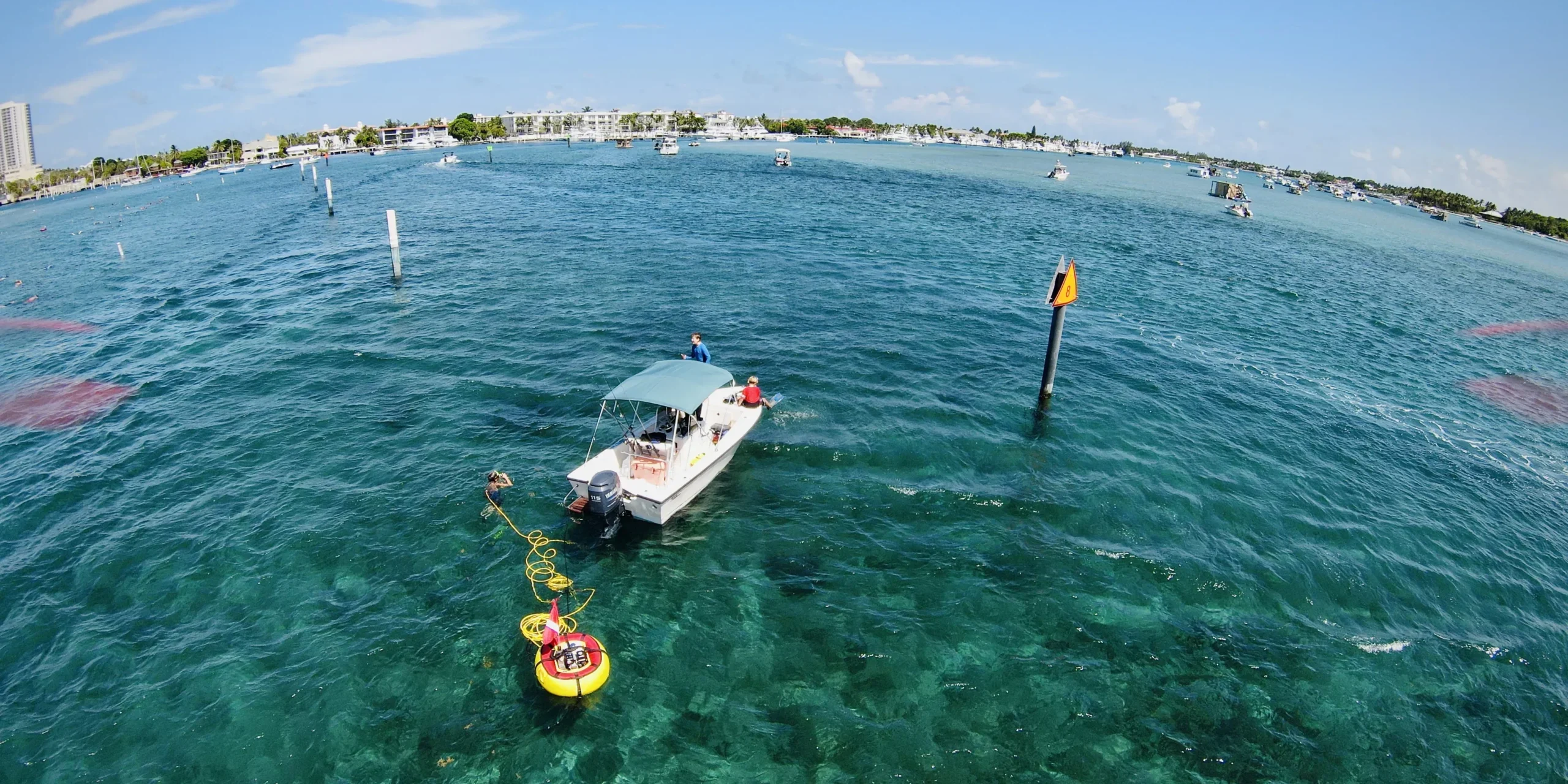 A diver uses a Brownie’s Third Lung tankless dive system connected to a small boat anchored in clear blue water near a channel marker, with the yellow and red floating compressor visible on the surface and other boats in the distance under a bright, sunny sky.