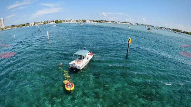A diver uses a Brownie&rsquo;s Third Lung tankless dive system connected to a small boat anchored in clear blue water near a channel marker, with the yellow and red floating compressor visible on the surface and other boats in the distance under a bright, sunny sky.