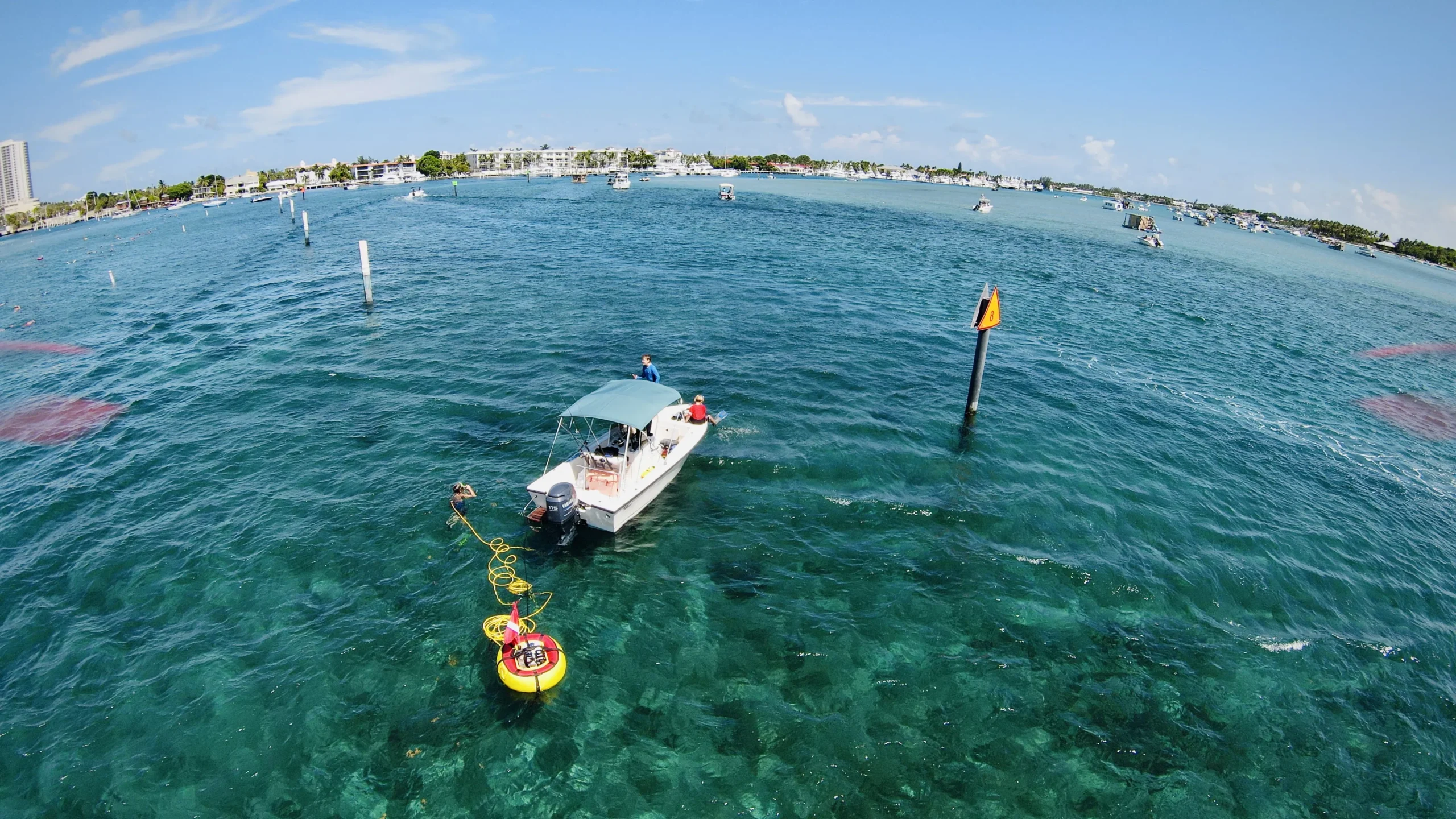 A diver uses a Brownie’s Third Lung tankless dive system connected to a small boat anchored in clear blue water near a channel marker, with the yellow and red floating compressor visible on the surface and other boats in the distance under a bright, sunny sky.
