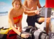 A woman and man prepare a Brownie’s Third Lung tankless dive system on a sandy beach, assembling the compressor and battery next to a red-and-yellow dive gear bag and a dive flag, with the ocean and pier visible in the background.