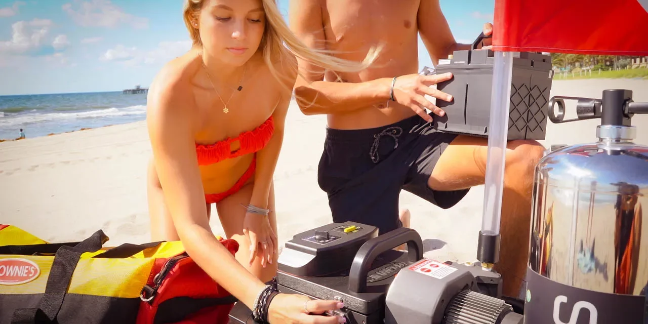 A woman and man prepare a Brownie&rsquo;s Third Lung tankless dive system on a sandy beach, assembling the compressor and battery next to a red-and-yellow dive gear bag and a dive flag, with the ocean and pier visible in the background.