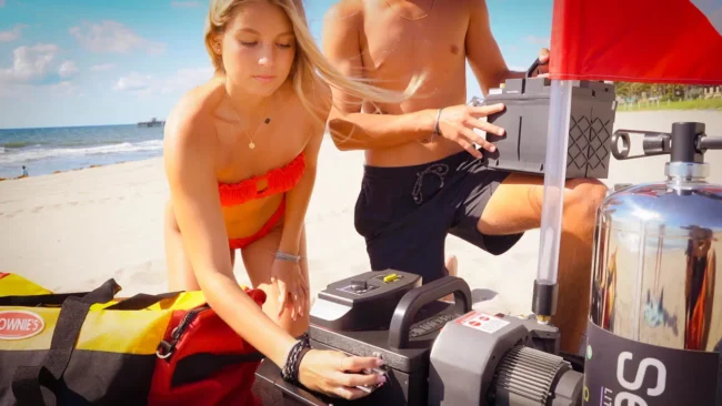 A woman and man prepare a Brownie&rsquo;s Third Lung tankless dive system on a sandy beach, assembling the compressor and battery next to a red-and-yellow dive gear bag and a dive flag, with the ocean and pier visible in the background.