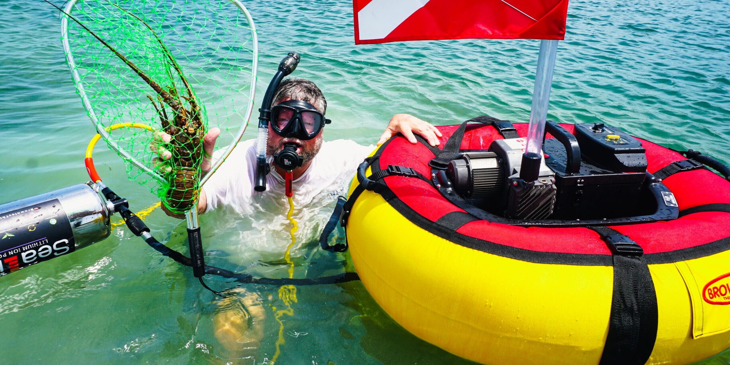 Diver using a Brownie’s Sea LiON floating hookah diving system to catch a lobster near the surface, with red and yellow float, dive flag, and battery-powered compressor.